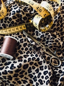 Detailed view of a sewing setup including a leopard print fabric, a yellow measuring tape, a spool of brown thread, and a pair of metal scissors.