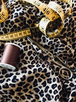 Detailed view of a sewing setup including a leopard print fabric, a yellow measuring tape, a spool of brown thread, and a pair of metal scissors.