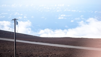 A lone power line stands on a barren landscape with rocky soil. A narrow paved road intersects the scene. Above, white clouds float against a backdrop of bright blue sky, giving the impression of a high elevation or mountaintop.
