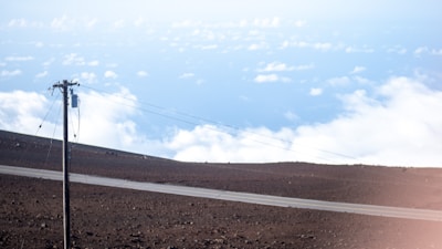 A lone power line stands on a barren landscape with rocky soil. A narrow paved road intersects the scene. Above, white clouds float against a backdrop of bright blue sky, giving the impression of a high elevation or mountaintop.