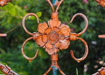 An ornate metal decoration with a floral pattern is prominently displayed. The intricate design features a flower in the center with petal-like elements and curving hooks extending outwards. The metal has a weathered, rusty appearance, contrasted against a lush, green, out-of-focus background.