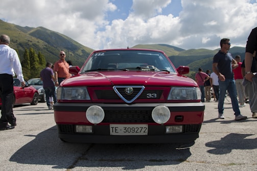 A classic red car with a license plate number TE-309272 is parked in an outdoor setting surrounded by people. The background features scenic hills and partly cloudy skies.