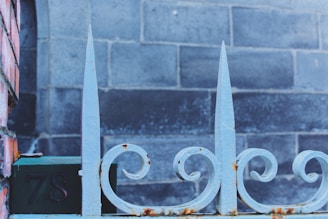 A close-up view of a decorative wrought iron fence with pointed tips and a floral swirl design. The fence has patches of rust and is set against a background of weathered gray bricks. The number '78' is visible on a dark plaque attached to the fence.