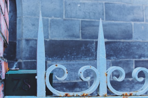 A close-up view of a decorative wrought iron fence with pointed tips and a floral swirl design. The fence has patches of rust and is set against a background of weathered gray bricks. The number '78' is visible on a dark plaque attached to the fence.