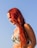 A person with vibrant red hair is wearing a patterned bikini top, with tropical designs of leaves and flowers. The backdrop is a clear blue sky, suggesting a sunny day.