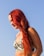 A person with vibrant red hair is wearing a patterned bikini top, with tropical designs of leaves and flowers. The backdrop is a clear blue sky, suggesting a sunny day.