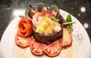 A beautifully arranged gourmet dish featuring a combination of seafood and steak. The centerpiece is a piece of seared steak topped with shrimp, mussels, and diced pineapple, garnished with herbs. Surrounding the steak are artfully sliced tomatoes and a rose-shaped tomato peel, all set on a white plate with a dark, elegant table surface.