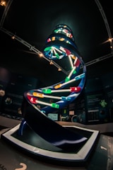 A large, illuminated DNA helix model is displayed in a darkened room, possibly an exhibit or a museum. The DNA model features colorful base pairs represented by differently colored bars with letters A, T, C, and G. The background includes informational posters and mounted lighting.