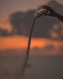 A silhouette of a hand holding a jar, pouring sand against a vibrant sunset backdrop with soft colors and a cloudy sky.