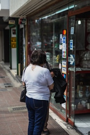 Two people are standing outside a shop with a glass display window. The window is adorned with various payment method stickers, including those for Visa and Mastercard. The individuals appear to be engaged in conversation, and one is wearing a white shirt and holding a black bag.