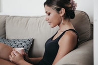 A woman sits comfortably on a beige couch, holding a pink mug. She has her hair tied back with a pink accessory and is dressed in a black tank top. A patterned cushion is beside her. Her posture and expression reflect a calm and relaxed demeanor.
