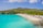 A picturesque beach scene featuring clear turquoise waters and a white sandy shore. People are enjoying various beach activities, including swimming and sunbathing. The shoreline is dotted with yellow sun loungers with thatched umbrellas, all set against a backdrop of lush green hills and a partly cloudy sky.