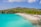 A picturesque beach scene featuring clear turquoise waters and a white sandy shore. People are enjoying various beach activities, including swimming and sunbathing. The shoreline is dotted with yellow sun loungers with thatched umbrellas, all set against a backdrop of lush green hills and a partly cloudy sky.