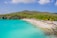 A picturesque beach scene featuring clear turquoise waters and a white sandy shore. People are enjoying various beach activities, including swimming and sunbathing. The shoreline is dotted with yellow sun loungers with thatched umbrellas, all set against a backdrop of lush green hills and a partly cloudy sky.