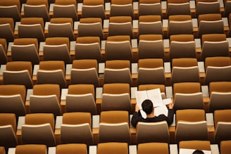 A lone person is seated in a large, empty auditorium. They are reading or reviewing sheet music at a desk surrounded by rows of empty, brown and beige seats.