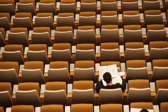 A lone person is seated in a large, empty auditorium. They are reading or reviewing sheet music at a desk surrounded by rows of empty, brown and beige seats.