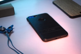A smartphone with a black leather case lies on a light surface, partially illuminated by soft lighting. Next to the phone, blue earphones with ear hooks are visible. A wooden phone case is also partially visible in the background, adding contrast and texture.
