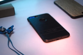 A smartphone with a black leather case lies on a light surface, partially illuminated by soft lighting. Next to the phone, blue earphones with ear hooks are visible. A wooden phone case is also partially visible in the background, adding contrast and texture.