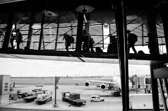 The image captures a scene at an airport with silhouetted figures walking through a glass corridor above. Below, an airplane is parked at the gate, with various service vehicles and equipment positioned around it. The tarmac extends into the distance, with a hint of a city skyline visible on the horizon.