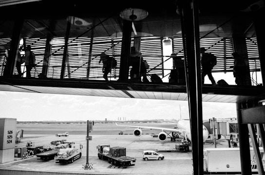 The image captures a scene at an airport with silhouetted figures walking through a glass corridor above. Below, an airplane is parked at the gate, with various service vehicles and equipment positioned around it. The tarmac extends into the distance, with a hint of a city skyline visible on the horizon.