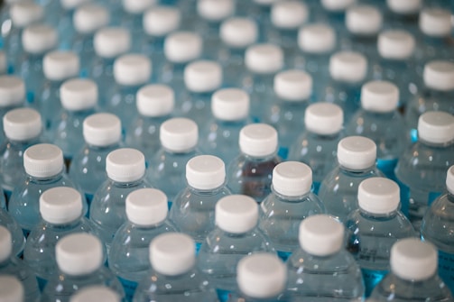A collection of clear plastic water bottles with white caps, tightly packed together and filled with light blue liquid. The bottles are arranged in a grid-like pattern.
