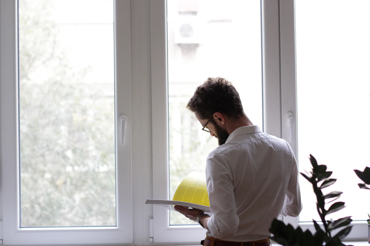 A site lead in a white shirt stands by a tall window reading through a binder, a yellow hard hat in hand.