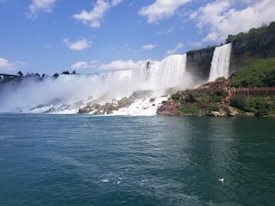 A majestic waterfall cascading over a cliff into a large body of water, with mist rising above and surrounding rocks. Lush greenery and wooden observation platforms are visible, with tourists walking along them. The sky is blue, scattered with fluffy clouds, creating a serene and powerful atmosphere.
