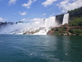 A majestic waterfall cascading over a cliff into a large body of water, with mist rising above and surrounding rocks. Lush greenery and wooden observation platforms are visible, with tourists walking along them. The sky is blue, scattered with fluffy clouds, creating a serene and powerful atmosphere.