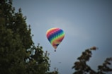 A vibrant multicolored hot air balloon is floating in the sky, with trees framing the sides of the image. The sky is overcast, providing a muted background that makes the bright colors of the balloon stand out.
