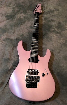 A sleek, pink electric guitar with a glossy finish is positioned on a textured gray background. It features a double cutaway body, black pickups, and tuning pegs. The strings and control knobs are clearly visible, with detailed craftsmanship on the bridge and fretboard.
