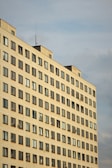 A high-rise residential building with rows of uniformly placed windows, some featuring satellite dishes. The structure's facade is a light, neutral color, and it's set against a cloudy sky.