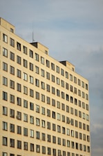 A high-rise residential building with rows of uniformly placed windows, some featuring satellite dishes. The structure's facade is a light, neutral color, and it's set against a cloudy sky.