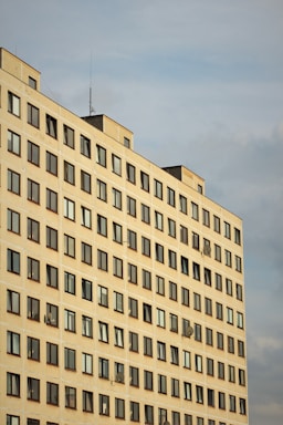 A high-rise residential building with rows of uniformly placed windows, some featuring satellite dishes. The structure's facade is a light, neutral color, and it's set against a cloudy sky.