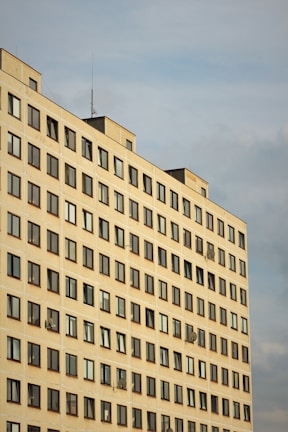 A high-rise residential building with rows of uniformly placed windows, some featuring satellite dishes. The structure's facade is a light, neutral color, and it's set against a cloudy sky.