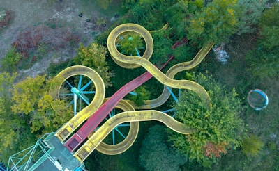An aerial view of a complex water slide structure with intertwining yellow slides surrounded by dense green foliage. The slides are supported by a blue metal framework and appear to be part of an amusement park or a water park. The vibrant colors of the slides contrast with the natural greenery of the trees and shrubs around them.