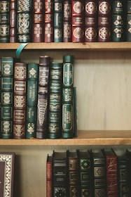 A collection of classic hardcover books with intricate gold designs and lettering on their spines. The books are tightly arranged on wooden shelves, showcasing a variety of titles and authors. The spines are predominantly green, blue, and burgundy, with ornate decorative patterns.