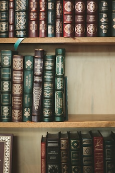 A collection of classic hardcover books with intricate gold designs and lettering on their spines. The books are tightly arranged on wooden shelves, showcasing a variety of titles and authors. The spines are predominantly green, blue, and burgundy, with ornate decorative patterns.