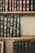 A collection of classic hardcover books with intricate gold designs and lettering on their spines. The books are tightly arranged on wooden shelves, showcasing a variety of titles and authors. The spines are predominantly green, blue, and burgundy, with ornate decorative patterns.
