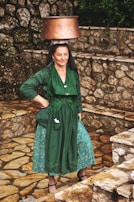 A woman is balancing a large copper pot on her head. She is wearing traditional attire including a green dress with intricate embroidery and a patterned blue skirt. The background consists of rustic stone walls and steps, creating a pastoral setting.