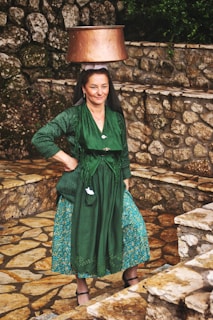 A woman is balancing a large copper pot on her head. She is wearing traditional attire including a green dress with intricate embroidery and a patterned blue skirt. The background consists of rustic stone walls and steps, creating a pastoral setting.