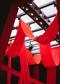 Red drapes hang elegantly from a metal frame, creating a dramatic and theatrical atmosphere. The structure features black metal beams and circular patterns that add a sense of industrial design against the vibrant fabric backdrop.