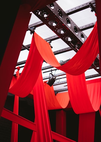 Red drapes hang elegantly from a metal frame, creating a dramatic and theatrical atmosphere. The structure features black metal beams and circular patterns that add a sense of industrial design against the vibrant fabric backdrop.