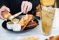 A person's hand is seen spreading green pesto onto a piece of toasted pita bread using a knife. The pita bread is served on a black plate alongside three small bowls filled with different dips. In the background, there is a glass of iced tea with a slice of lemon.