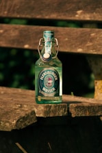 A glass bottle of Flensburger Wasser mineral water is placed on a rustic wooden bench outdoors. The bottle has a swing-top cap and a label featuring a ship design. The sunlight casts shadows and highlights the textures of both the bottle and the weathered wood.