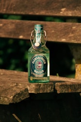 A glass bottle of Flensburger Wasser mineral water is placed on a rustic wooden bench outdoors. The bottle has a swing-top cap and a label featuring a ship design. The sunlight casts shadows and highlights the textures of both the bottle and the weathered wood.