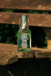A glass bottle of Flensburger Wasser mineral water is placed on a rustic wooden bench outdoors. The bottle has a swing-top cap and a label featuring a ship design. The sunlight casts shadows and highlights the textures of both the bottle and the weathered wood.