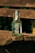 A glass bottle of Flensburger Wasser mineral water is placed on a rustic wooden bench outdoors. The bottle has a swing-top cap and a label featuring a ship design. The sunlight casts shadows and highlights the textures of both the bottle and the weathered wood.