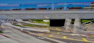 A digitally altered or distorted view of a highway intersection with multiple lanes and bright yellow lines. Overhead, there is a large concrete bridge or overpass with a train visible on top. The scene includes sections of road markings, concrete barriers, grassy areas, and parts of the infrastructure appear misaligned, creating a surreal effect.