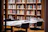 A library scene with shelves filled with a variety of books. In the foreground, a table is covered with stacked books and papers, surrounded by modern black chairs. The shelves are made of wood and add a warm, inviting feel to the room.