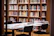 A library scene with shelves filled with a variety of books. In the foreground, a table is covered with stacked books and papers, surrounded by modern black chairs. The shelves are made of wood and add a warm, inviting feel to the room.